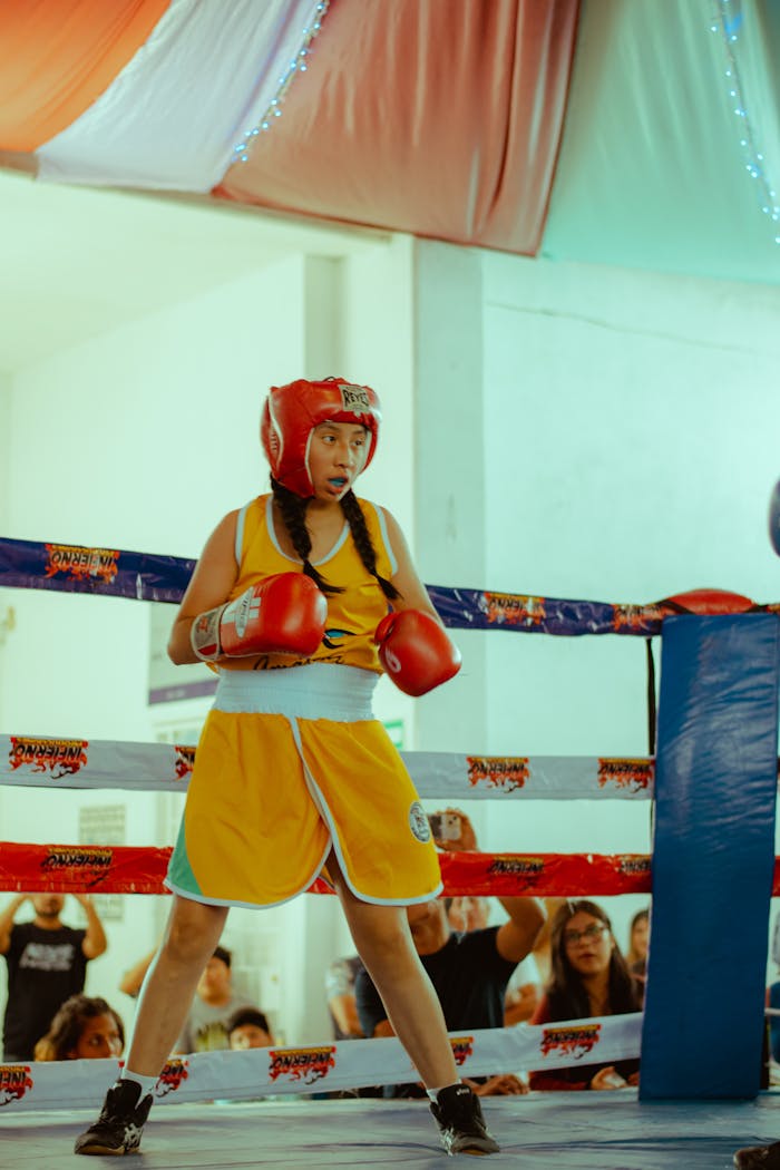 A young female boxer dressed in red headgear and yellow shorts stands in a boxing ring, ready for the match.